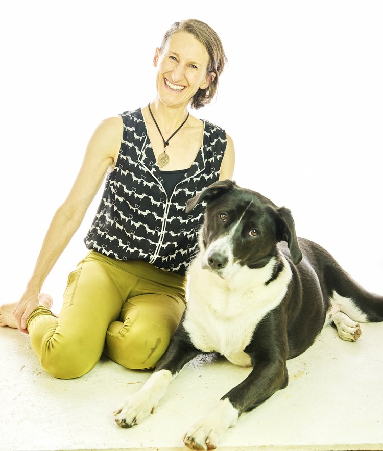 Image shows Sharon, smiling radiantly, in moss green leggings and sleeveless dog print top, seated on the floor next to Muggins, a black-and-white floppy-eared pup rests at her side. Words in forest green announce: I am on 2 podcasts!