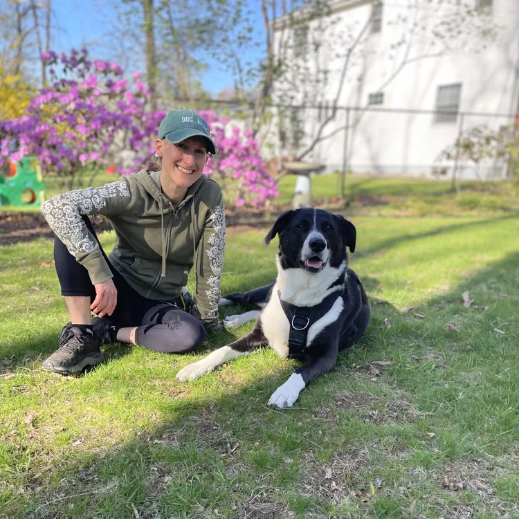 Image shows a large black-and-white dog laying in grass next to a seated white human wearing green hoodie and teal "dog mom" cap. The two smile as they lock eyes. Sunshine adorns yellow and pink blooms on bushes in the background.