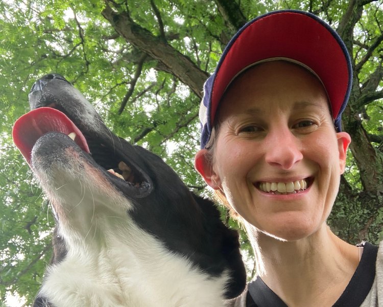 Image shows Sharon, a white human smiling radiantly at the camera while wearing a red and blue ball cap and standing under a glorious tree and next to Muggins, a happy black-and-white dog.