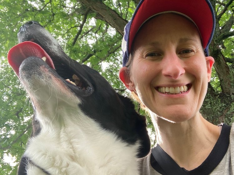 Image shows Sharon, a white human wearing a green leggings and black vest, sitting on the ground next to Muggins, a large black-and-white dog. The two connect in quiet eye contact and treat exchange.