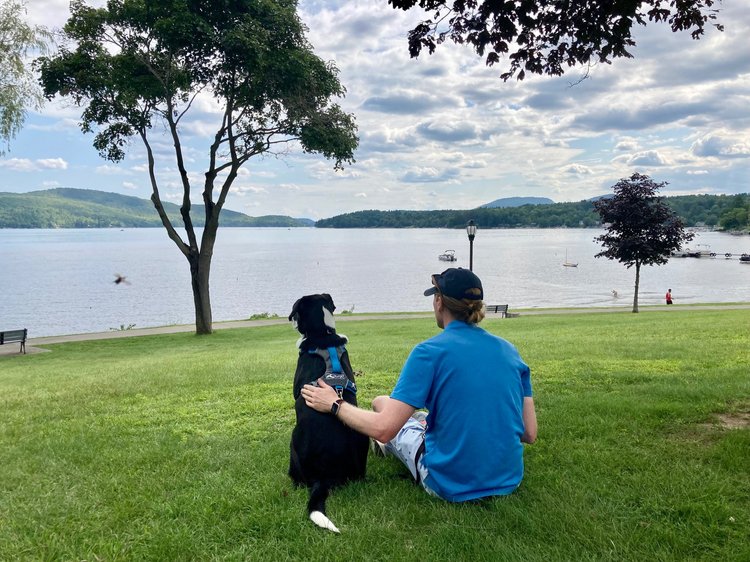 Image shows a human and a dog, seated next to each other on a grassy hill above an Adirondack lake, the sky is bursting with fluffy clouds as sun streams through the atmosphere. The human places a tender hand on the back of the dog.