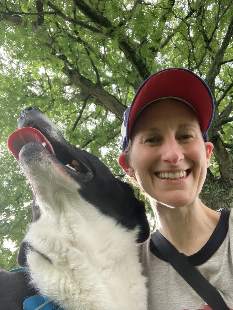 Image shows Sharon, a white human wearing a red and blue ca, and grey shirt, smiling radiantly next to Muggins, a large black-and-white dog smiling at the sun. The two are underneath a green canopy.