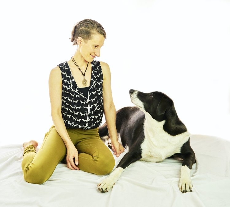 Image shows Sharon, seated in moss green leggings and sleeveless black-and-white dog print top, while catching eye contact with Muggins, a black-and-white floppy-eared pup who is laying by their side.