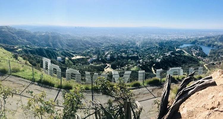 Image shows a mountain top view from behind the Hollywood sign. In the distance you can see the Verdugo mountains, Hollywood reservoir and silhouette of downtown Los Angeles.