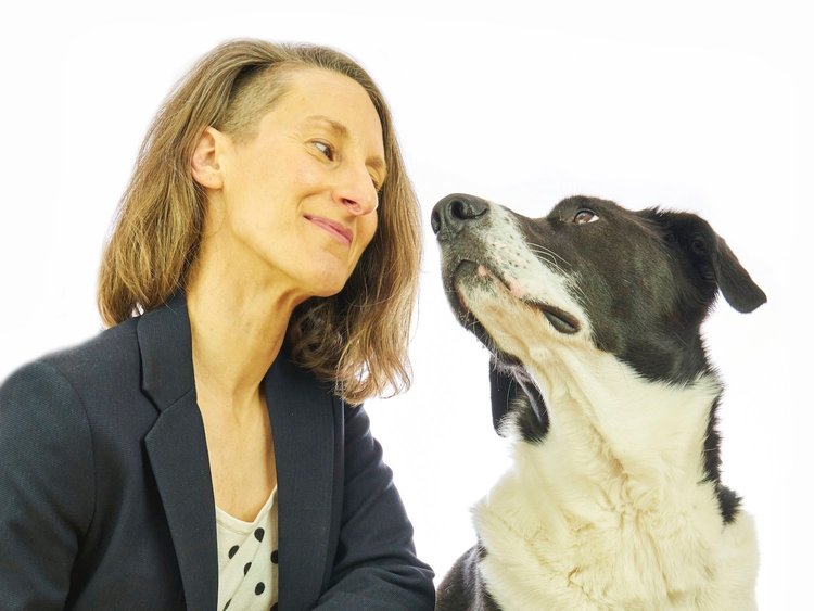 Image shows Sharon, a white human wearing shoulder-length brown hair and navy blazer, sitting beside Muggins, a black-and-white pup. The two look at each other smilingly.