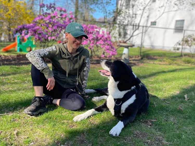 Image shows a large black-and-white dog laying in grass next to a seated white human wearing green hoodie and teal "dog mom" cap. The two smile as they lock eyes. Sunshine adorns yellow and pink blooms on bushes in the background.