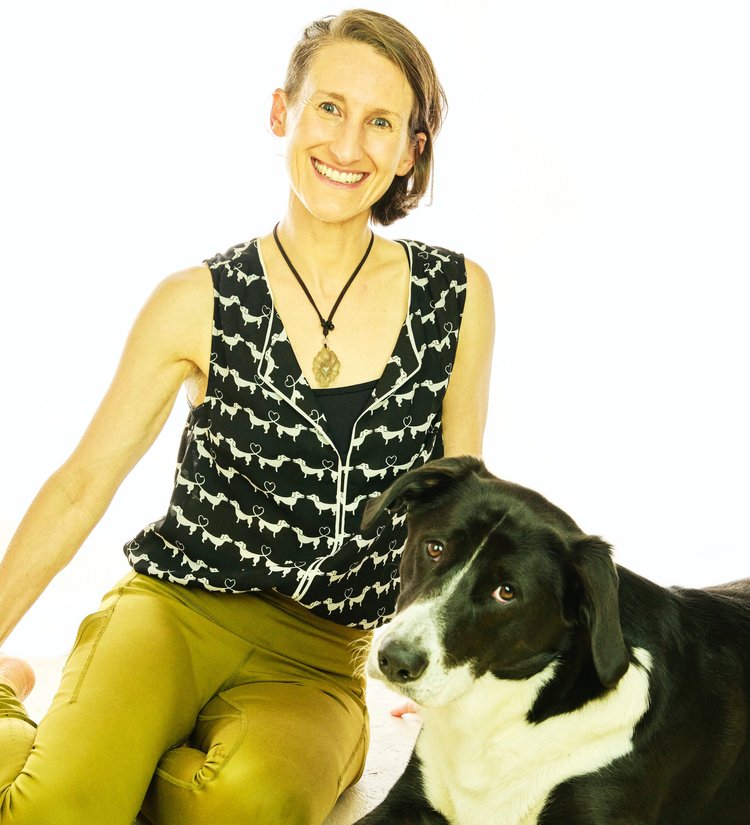 Image shows Sharon, a white human smiling radiantly and wearing olive leggings and black-and-white top, seated next to Muggins, a large floppy-eared black-and-white dog who is adorably looking at the camera. 