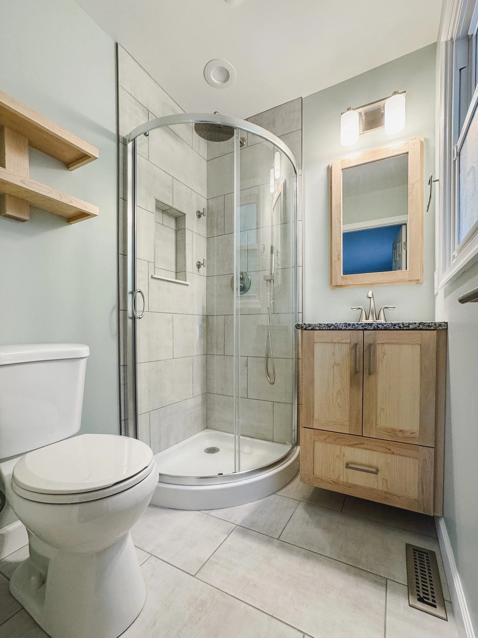Remodeled primary bathroom in Sykesville with curved glass shower, custom wood vanity, shelves, mirror frame, and new lighting