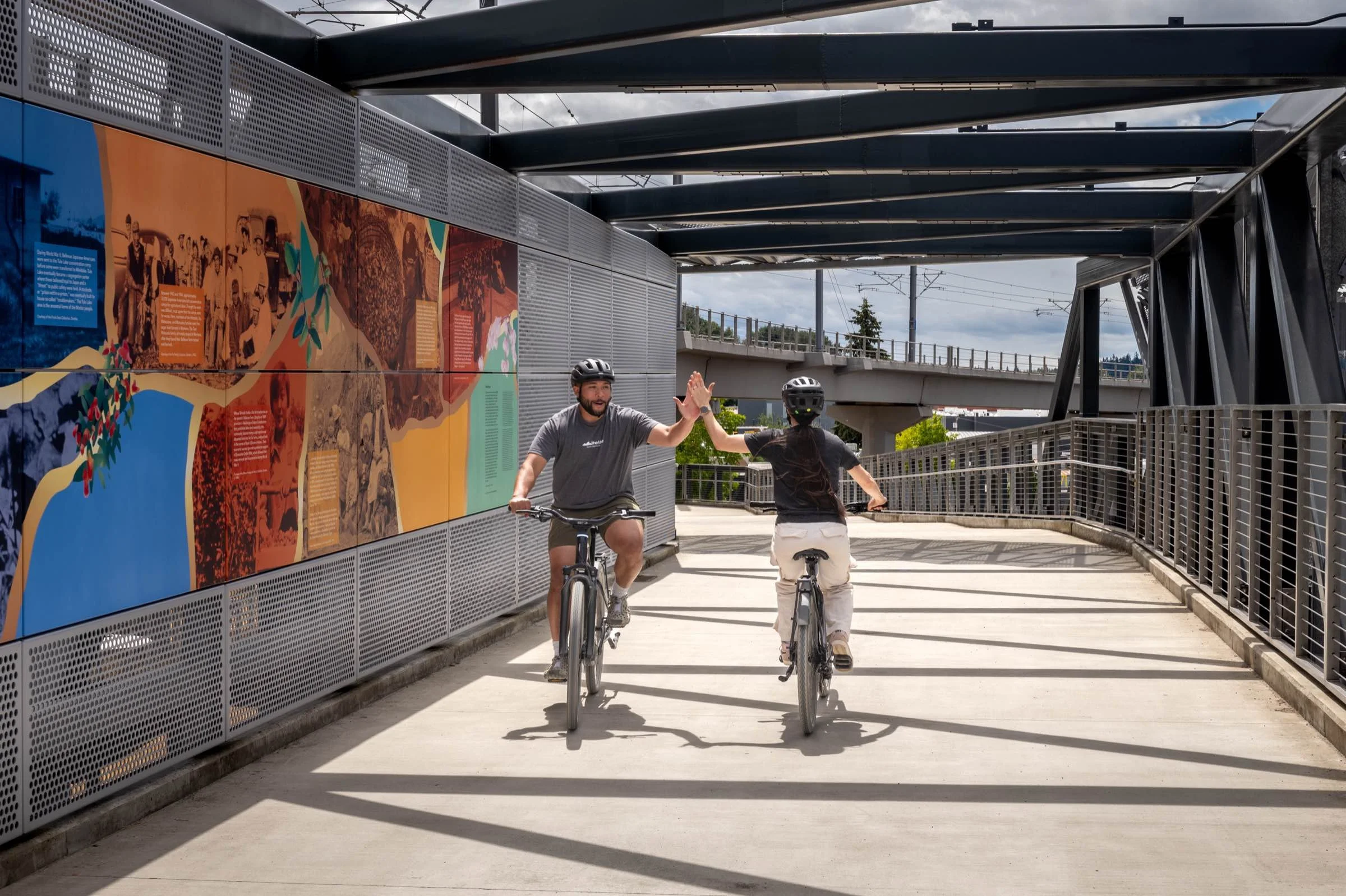 Two riders high-fiving as they ride the NE 8th Street bridge on the Eastrail, the art installation running along the wall beside them