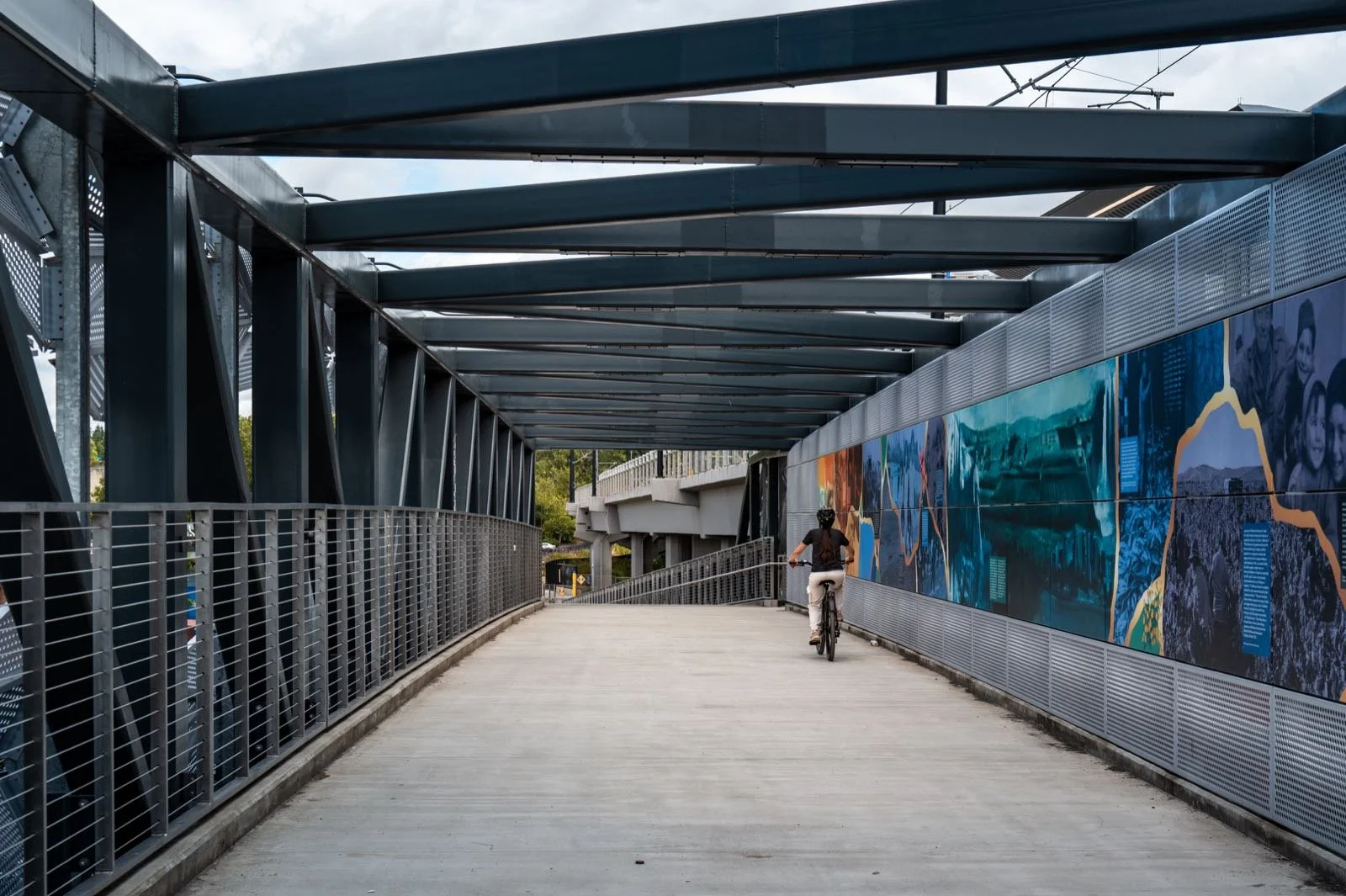 The NE 8th Street trail bridge seen down its length, steel structure overhead and the art and poetry panels running the full wall, a lone rider for scale