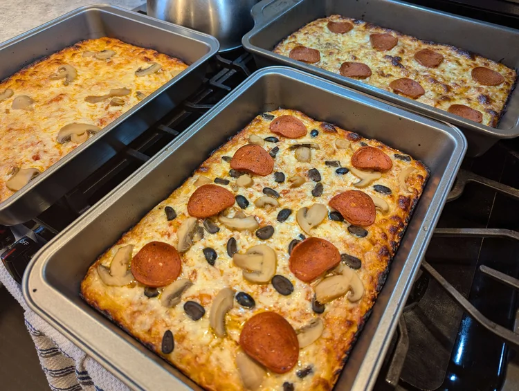 Three rectangular pan-pizzas in 9x13” metal pans on a stove top. The cheese has crusted and browned around the edges. One is mushroom, one is pepperoni, and one has mushroom, pepperoni, and black olives.