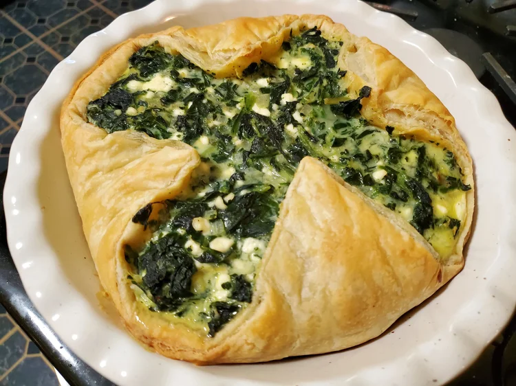 In a white pie plate, golden corners of puff pastry are folded up around a spinach and cheese filling. The exposed filling is in a green, plus-sign shape.