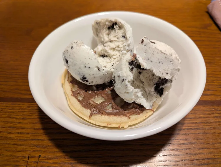A bowl with a round waffle at the bottom. A layer of chocolate-hazelnut spread outlines the waffle squares. Two curled scoops of cookies ‘n’ cream ice cream are on top of the waffle.