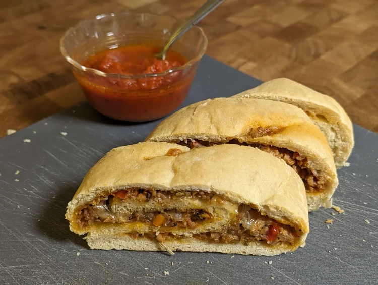 Three thick slices of stromboli on a gray cutting board, with a small glass dish of pizza sauce in the background. The spiral of one slice is towards the camera, with visible sausage crumbles, spices, and cheese wrapped in bread.