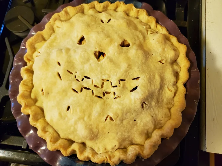 A double-crusted pie in a purple ceramic pie plate, viewed from the top. Vents have been cut into the top crust that resemble a jack-o-lantern face, triangles for eyes and nose, and a big smile with square teeth.