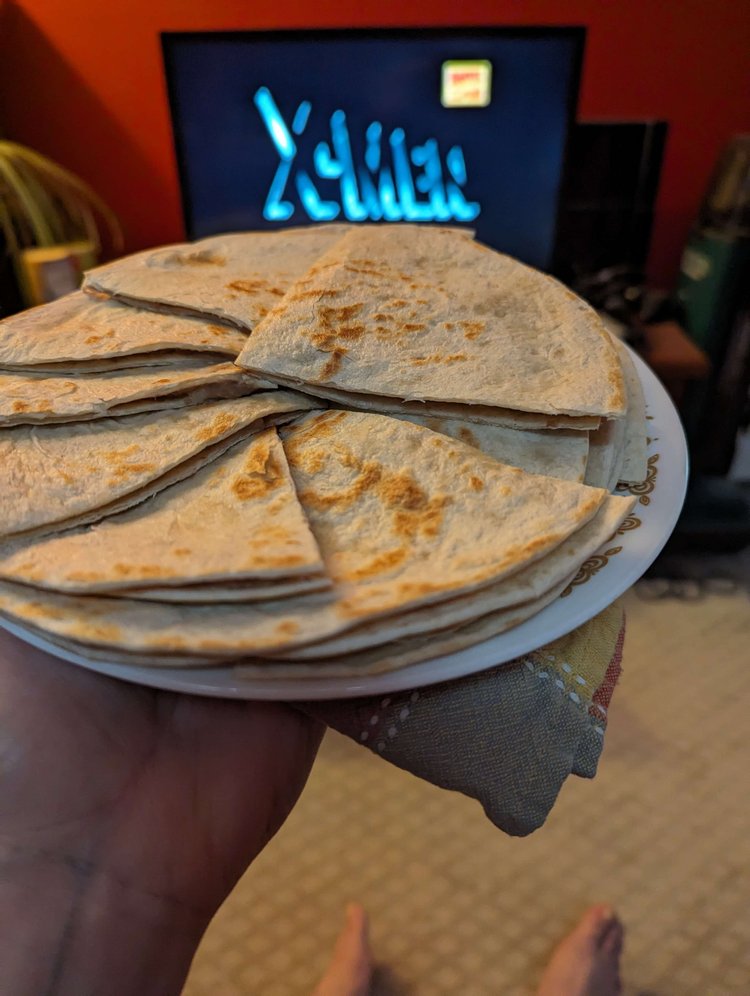 Jex (not pictured) is holding a plate of bean-and-cheese quesadillas cut in quarter-circles in their living room. In the blurred background above the plate is a TV screen with the X-Men logo.