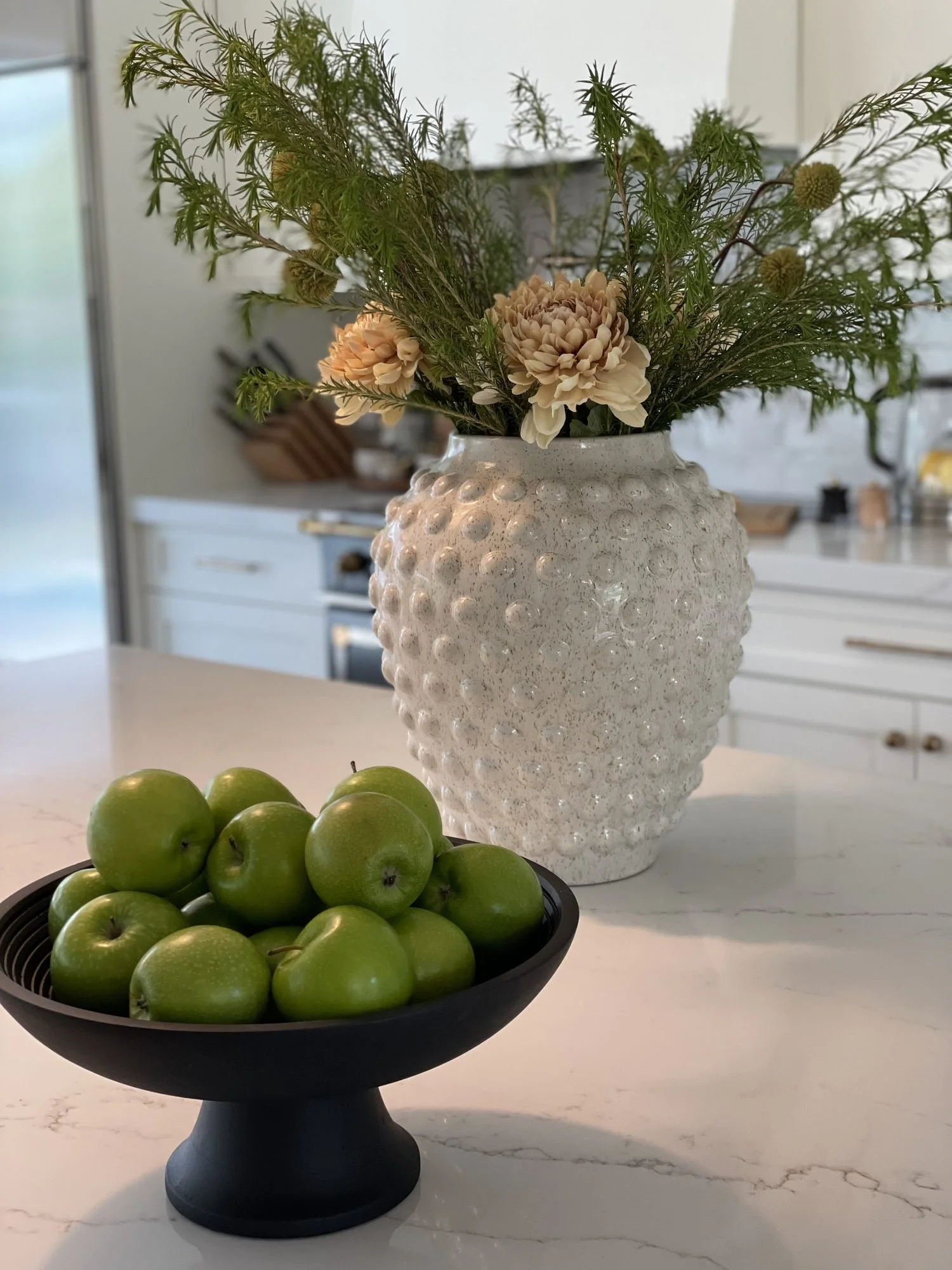 A hobnail vase and a bowl of green apples on a marble counter