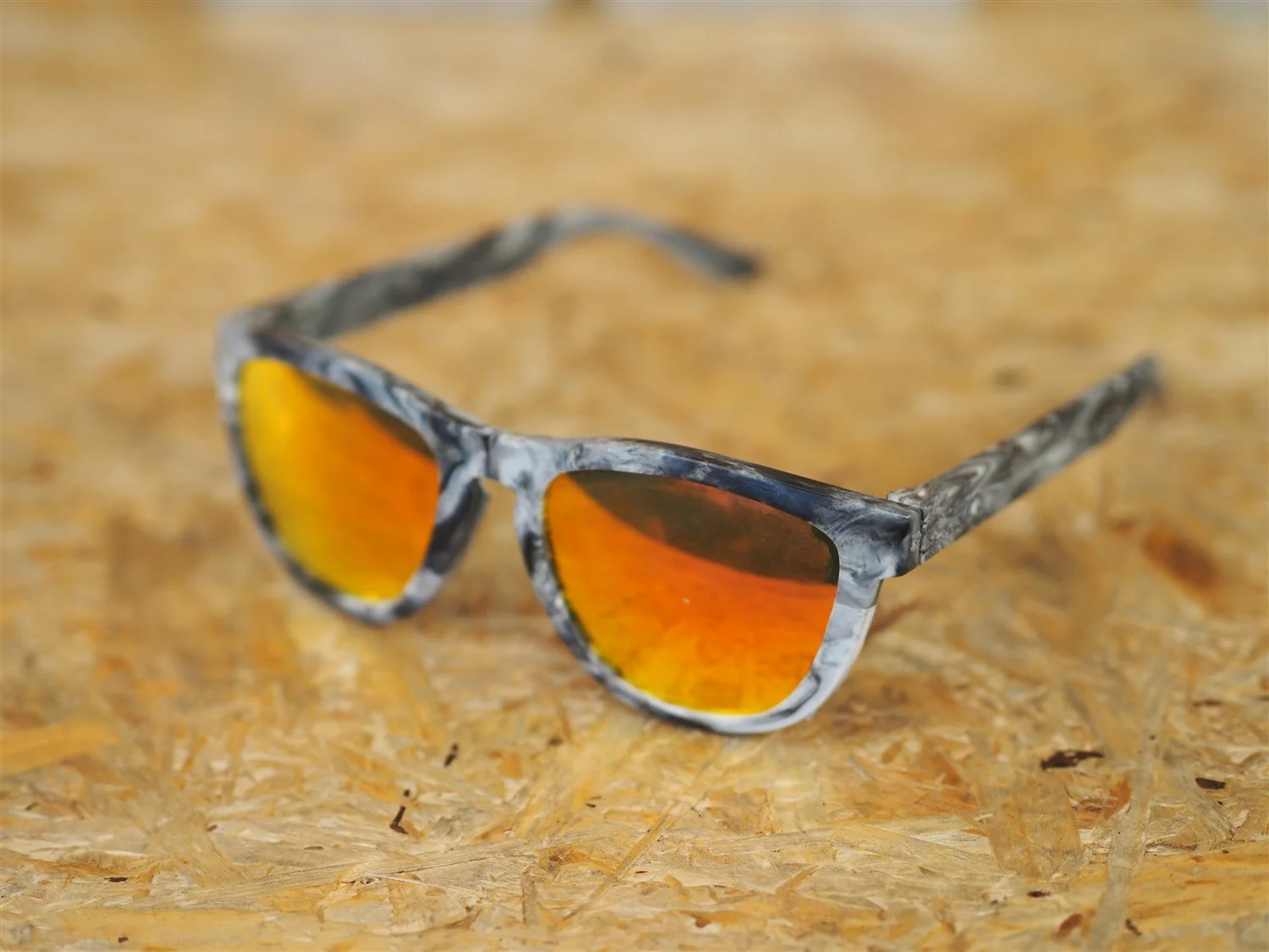The finished pair of recycled plastic sunglasses, marbled white, grey and black frame with orange mirrored lenses, arms open, resting on an OSB bench