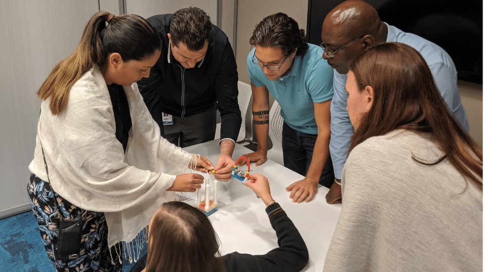 Six colleagues crowd around a small white table, leaning in as two of them add pieces to a shared LEGO model.