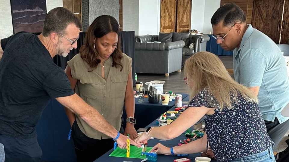 Four leaders stand around a table and reach in together to place pieces on one LEGO model.