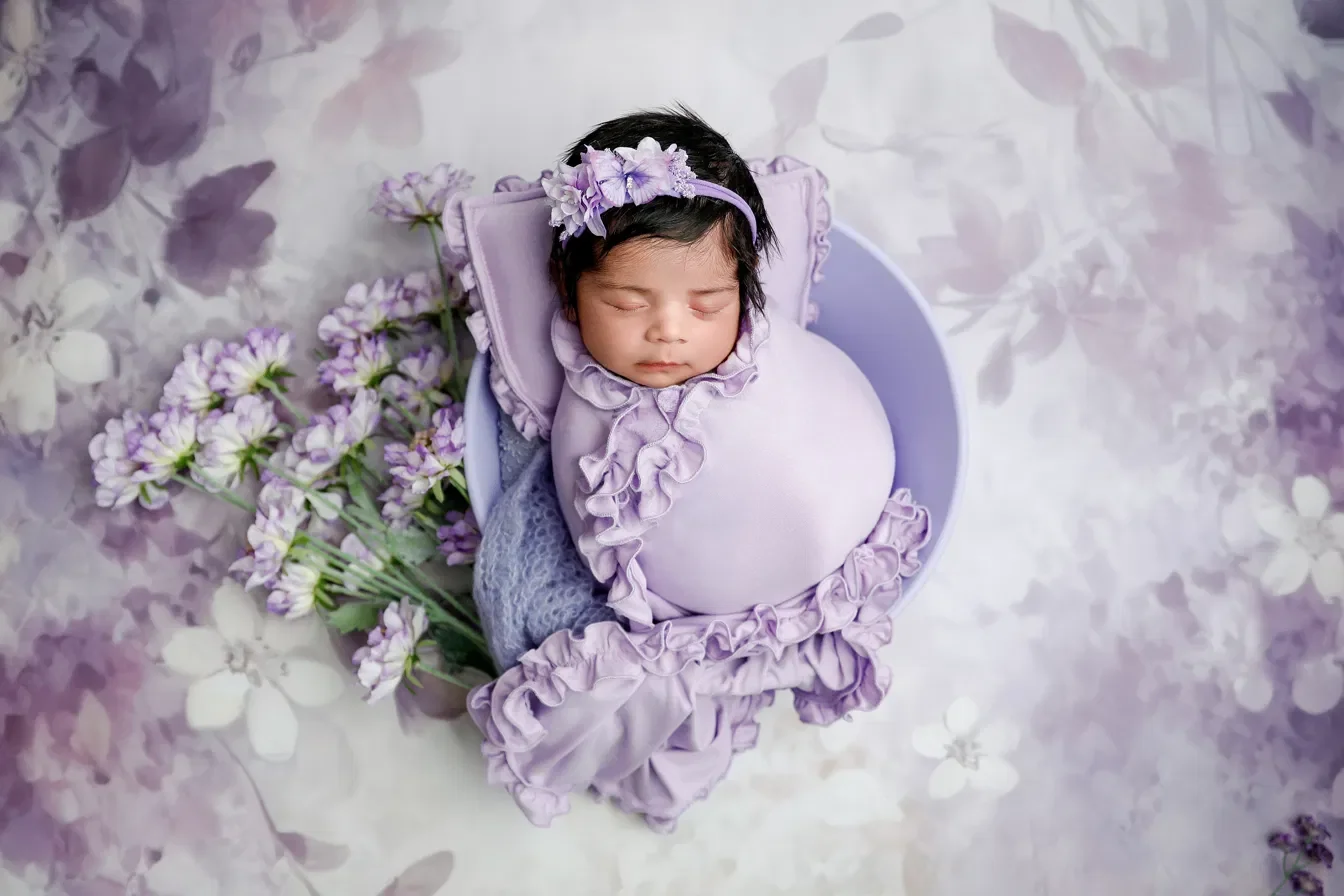 Newborn in a cream pom pom hat posed upright in a white bucket with a draped blanket