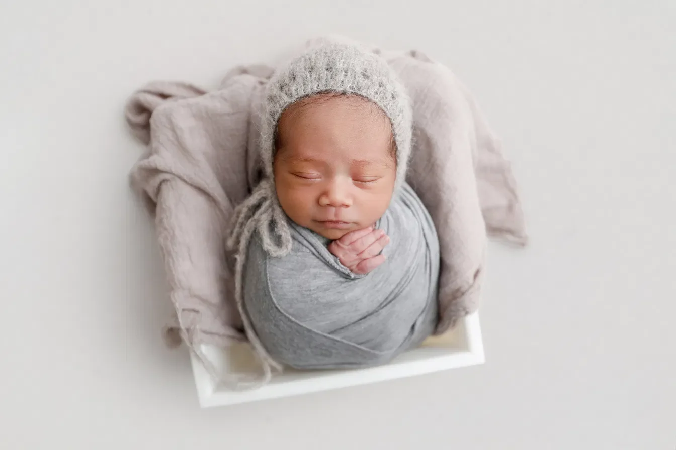 Newborn in a grey wrap and knit bonnet with one hand showing, posed in a small white crate