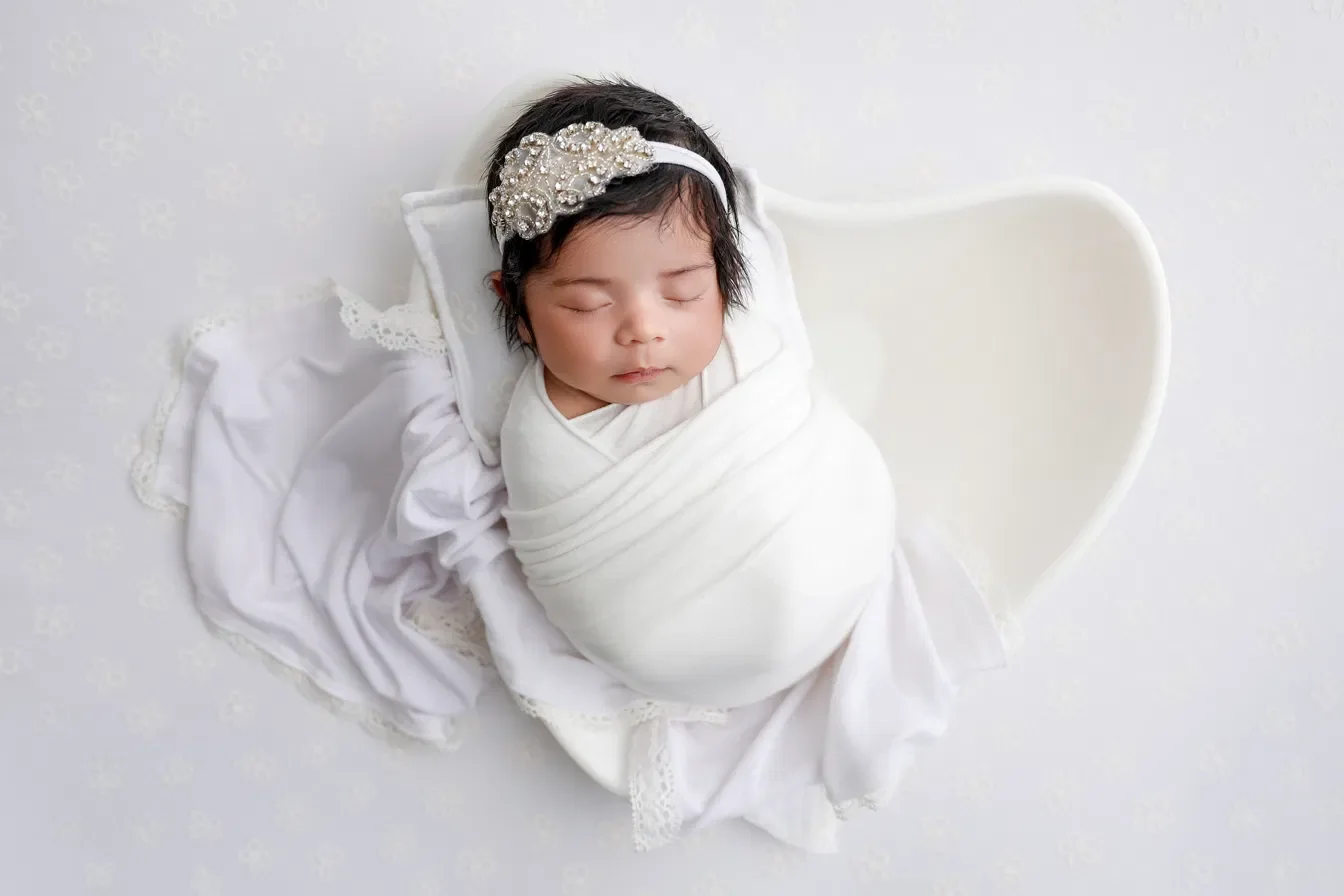Newborn girl wrapped in white with a rhinestone headband, posed in a white heart shaped bowl