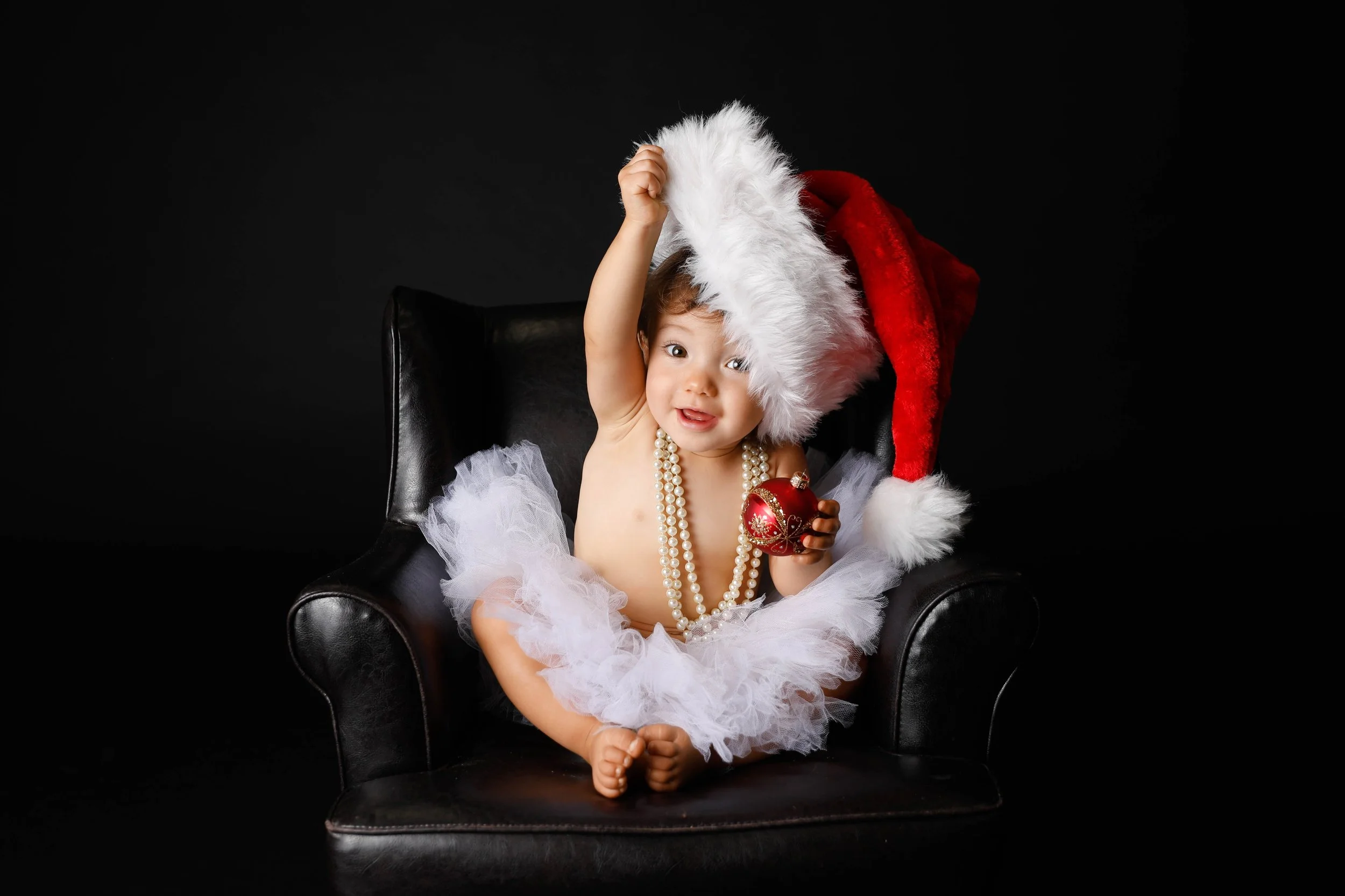 Baby in a Santa hat and white tutu holding a red ornament
