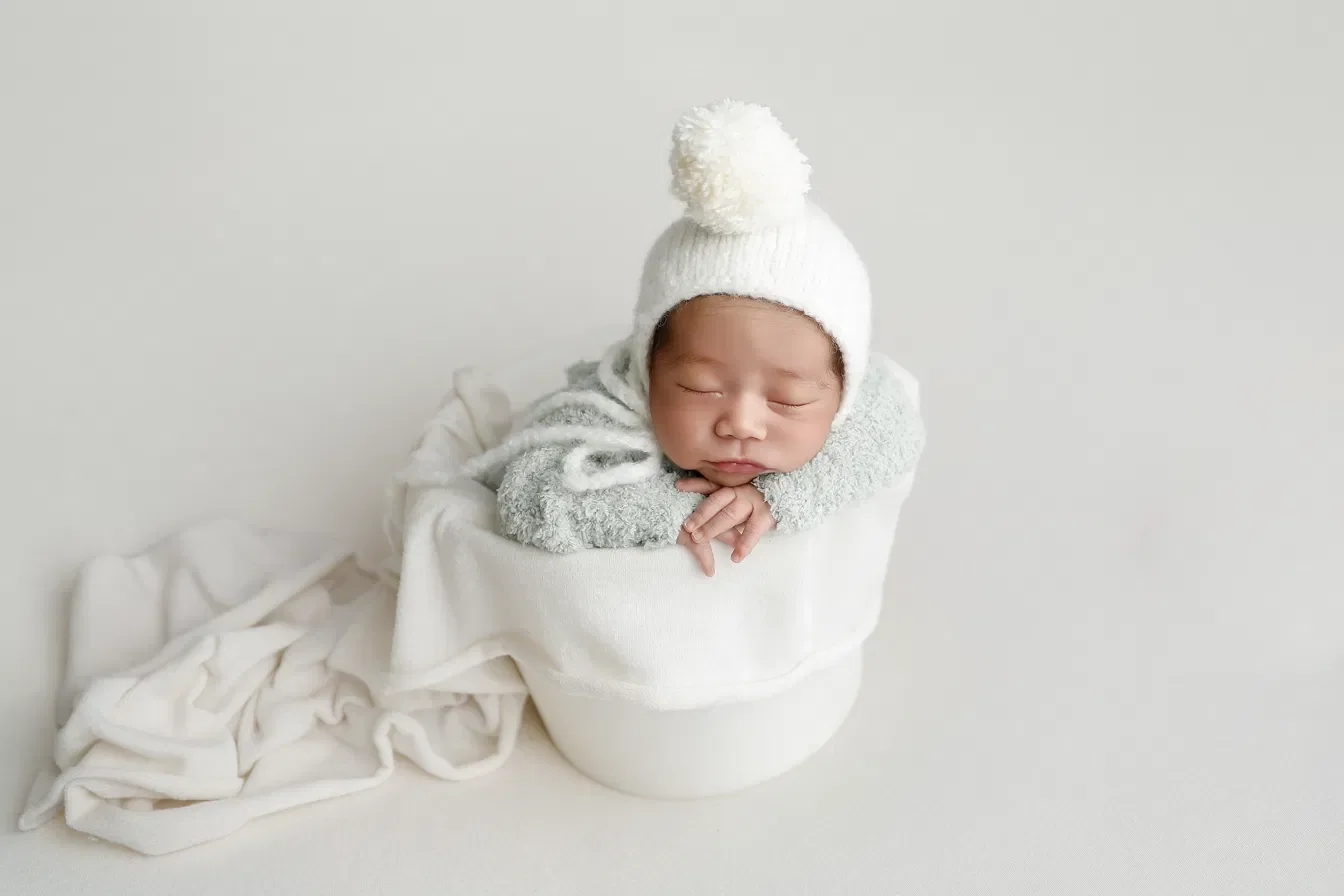 Newborn in a green bear bonnet resting hands on the rim of a white bucket, posed by Ana Brandt