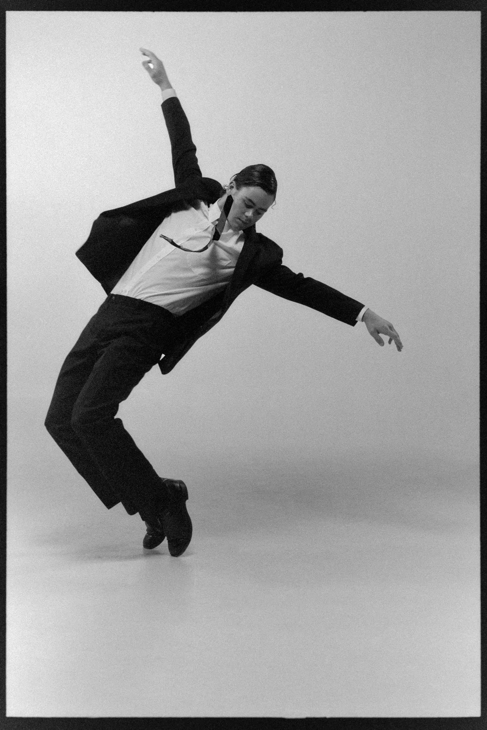 Dancer in a dark suit leaning back mid-movement, arm raised, in a white studio