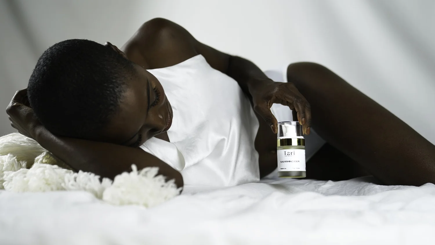 Editorial lifestyle skincare product photography of a model reclining on white bedding holding Tari serum, shot with soft directional light, controlled highlights and natural skin tones