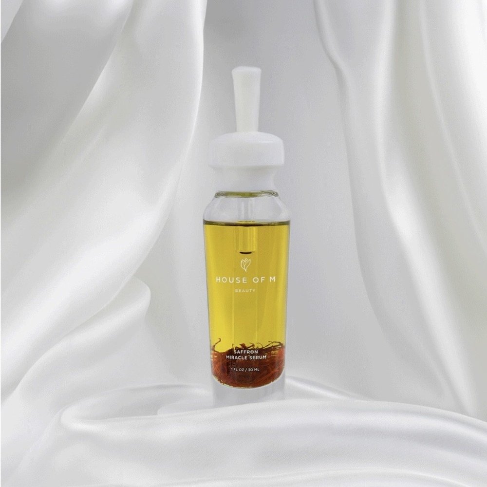 Golden House of M Beauty Saffron Miracle Serum framed by flowing white fabric, photographed in soft high-key light that reveals the translucent oil, saffron detail and glass packaging without distracting reflections