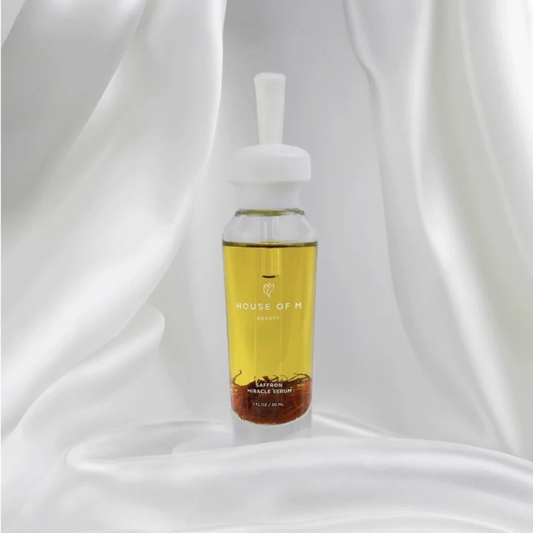 Golden House of M Beauty Saffron Miracle Serum framed by flowing white fabric, photographed in soft high-key light that reveals the translucent oil, saffron detail and glass packaging without distracting reflections