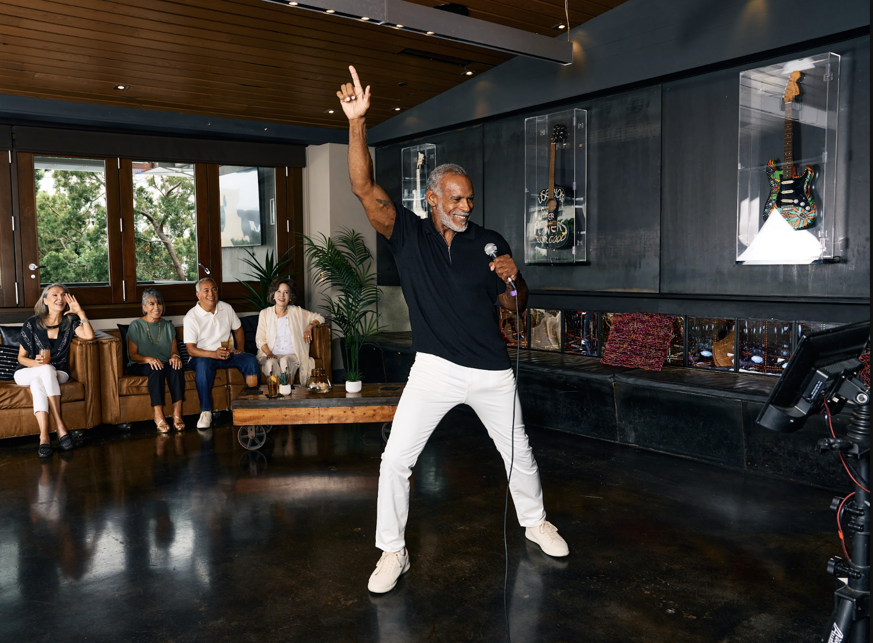 Older man in a black polo and white jeans singing karaoke with one arm raised while four friends cheer from a leather sofa in a lounge, photographed for an agency lifestyle campaign in San Diego County