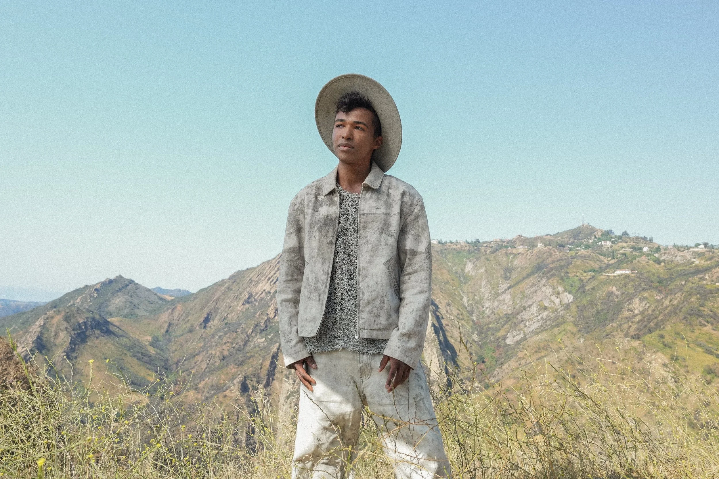 Menswear fashion photography: model in a washed canvas work jacket, open-knit top and wide-brim hat on a Southern California hillside