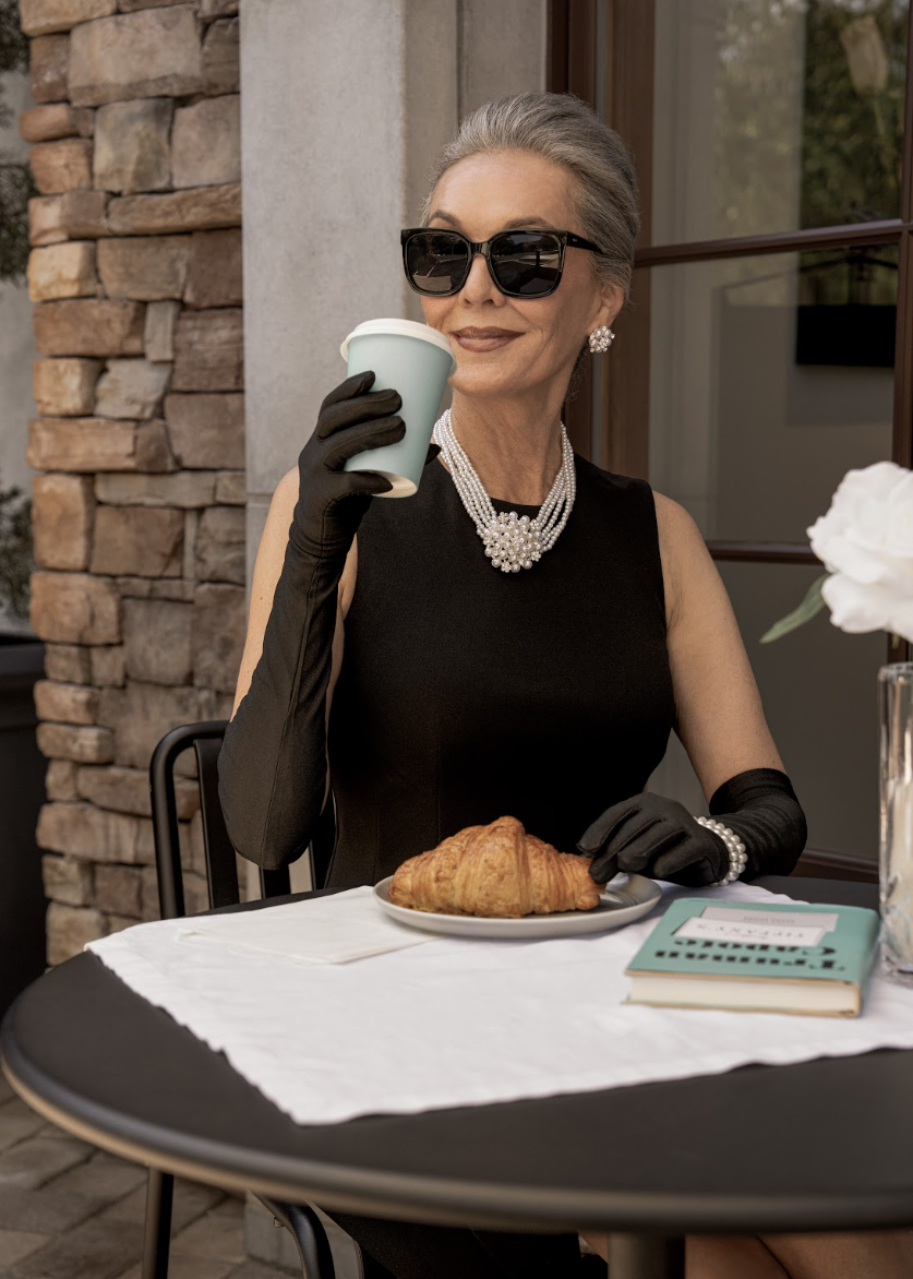 Silver-haired woman in a black dress, long gloves, pearls and sunglasses sipping coffee beside a croissant at an outdoor cafe table, styled for an agency lifestyle campaign in Southern California