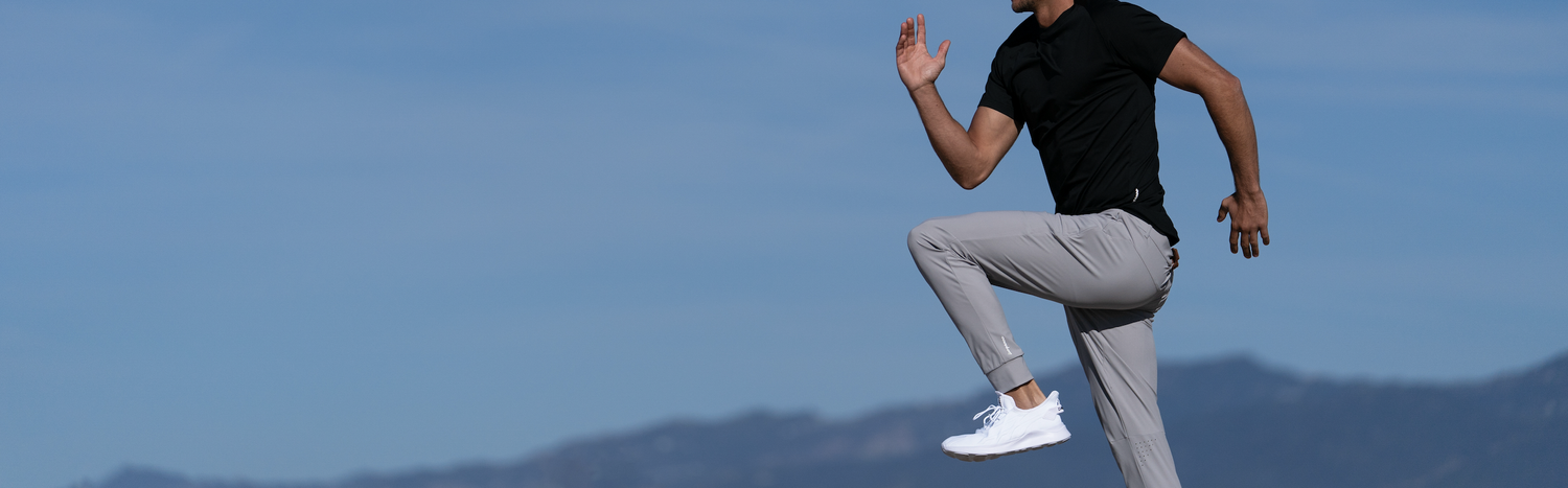 Commercial lifestyle photograph of a male athlete captured in a dynamic high-knee running motion, wearing black performance apparel and grey athletic pants in Los Angeles. Precise action timing, directional natural light and minimalist framing against a blue mountain backdrop create a striking, movement-driven image