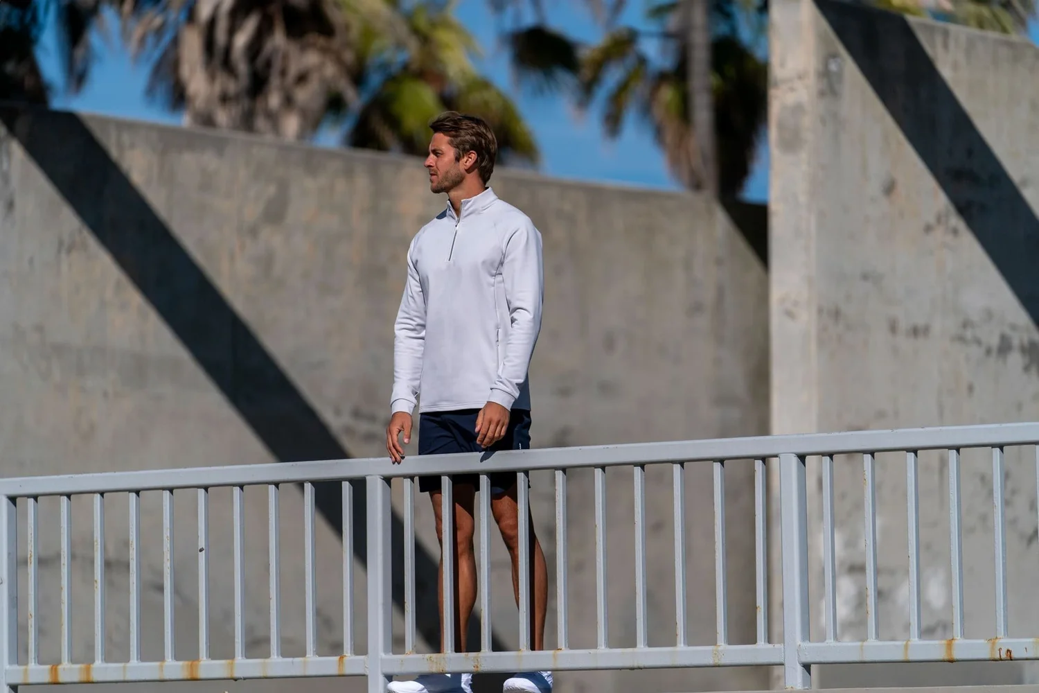 Premium men's activewear lifestyle photography in Venice Beach, Los Angeles, featuring a male model wearing a light grey quarter-zip performance top, navy athletic shorts and white sneakers against modern concrete architecture. Natural California sunlight highlights the apparel's fit, texture and design in this commercial sportswear campaign.