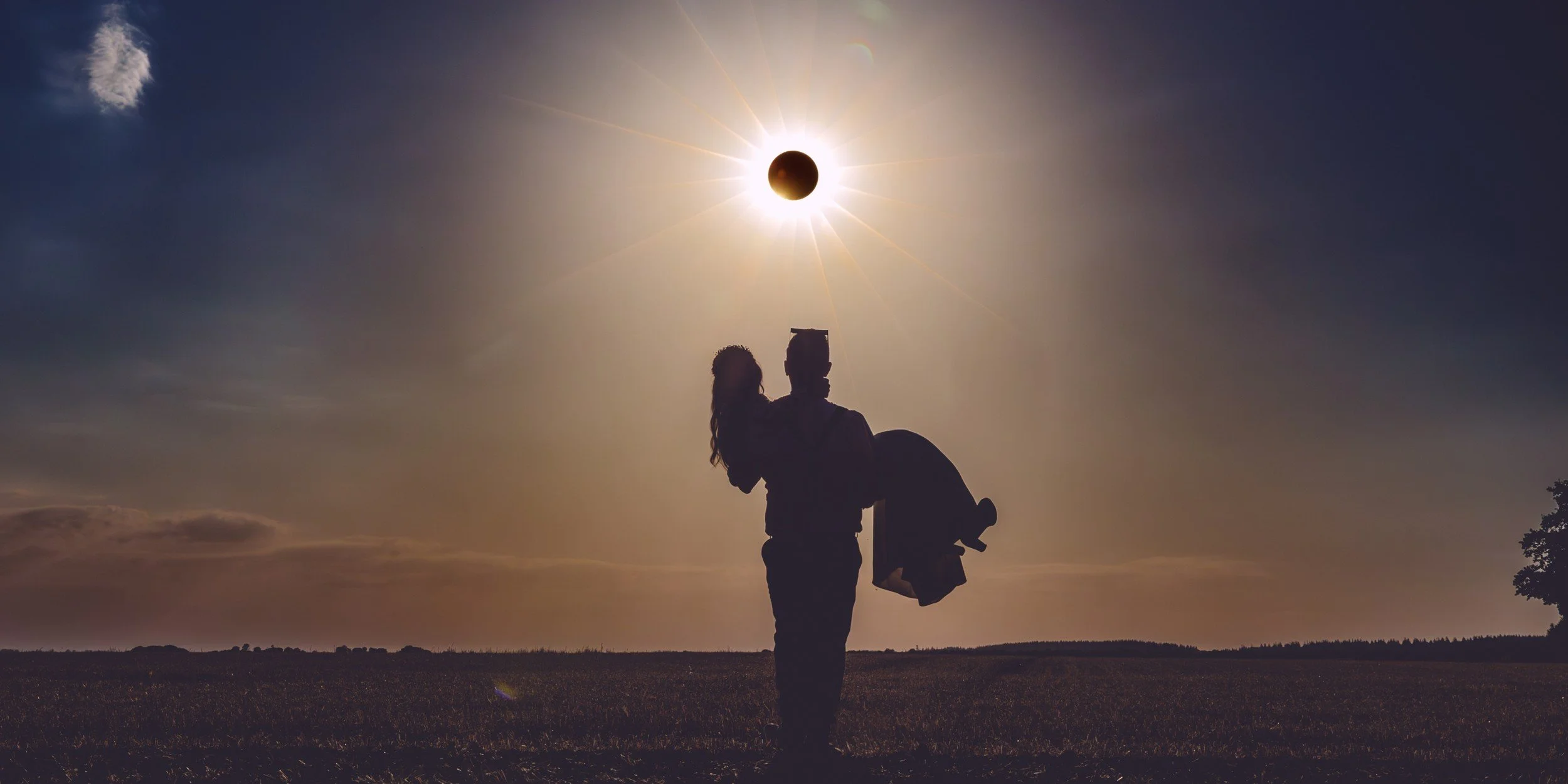 Groom carrying his bride beneath an eclipse-like sun, silhouetted in a field