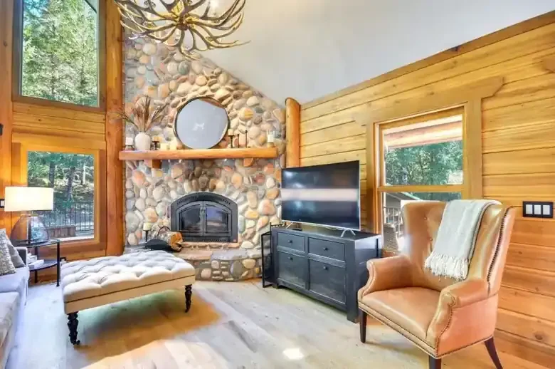 A cabin living room with a stone fireplace, comfortable seating and forest views