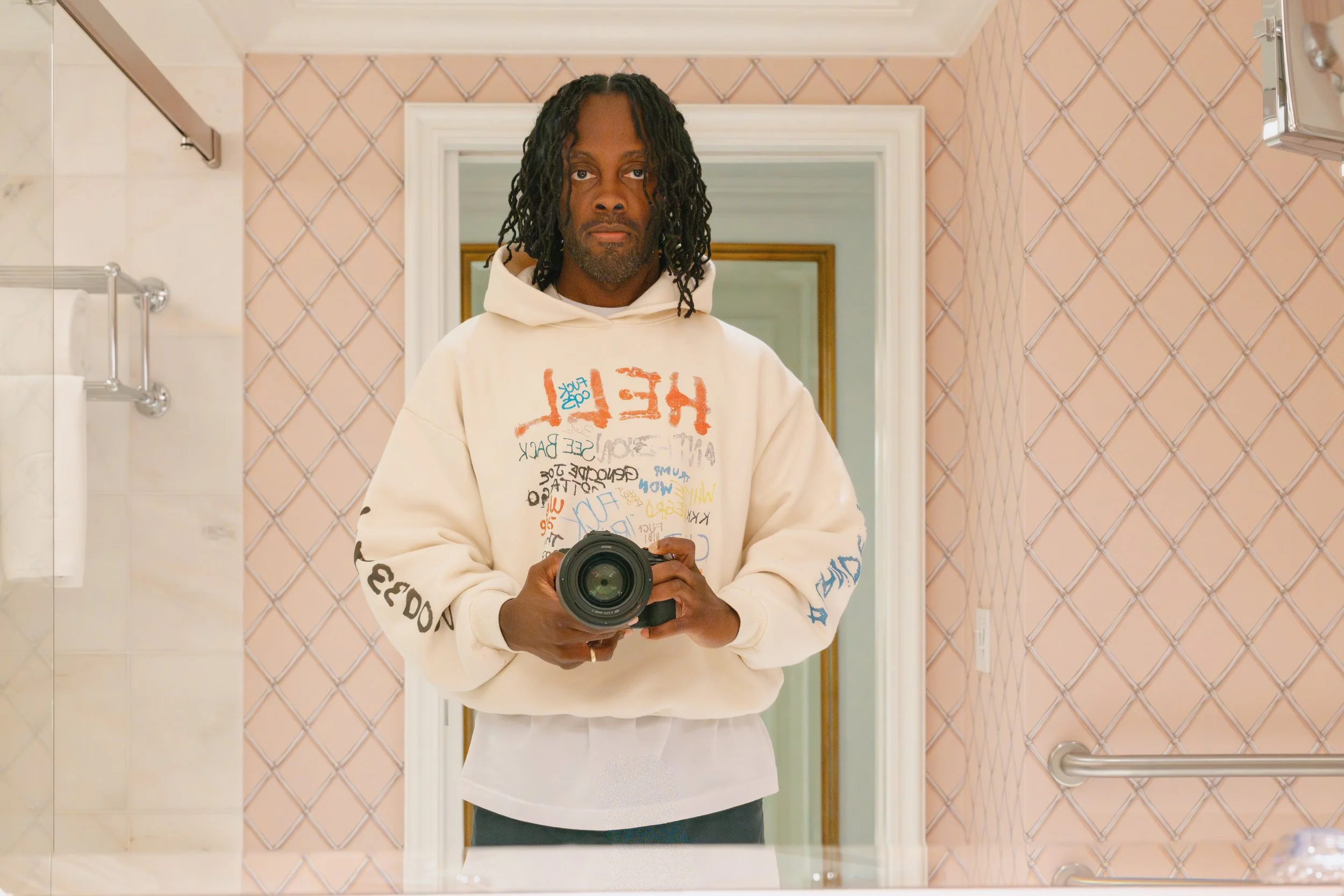 Self-portrait in a tiled bathroom wearing the HELL ON EARTH fieldwear garment while holding a camera.