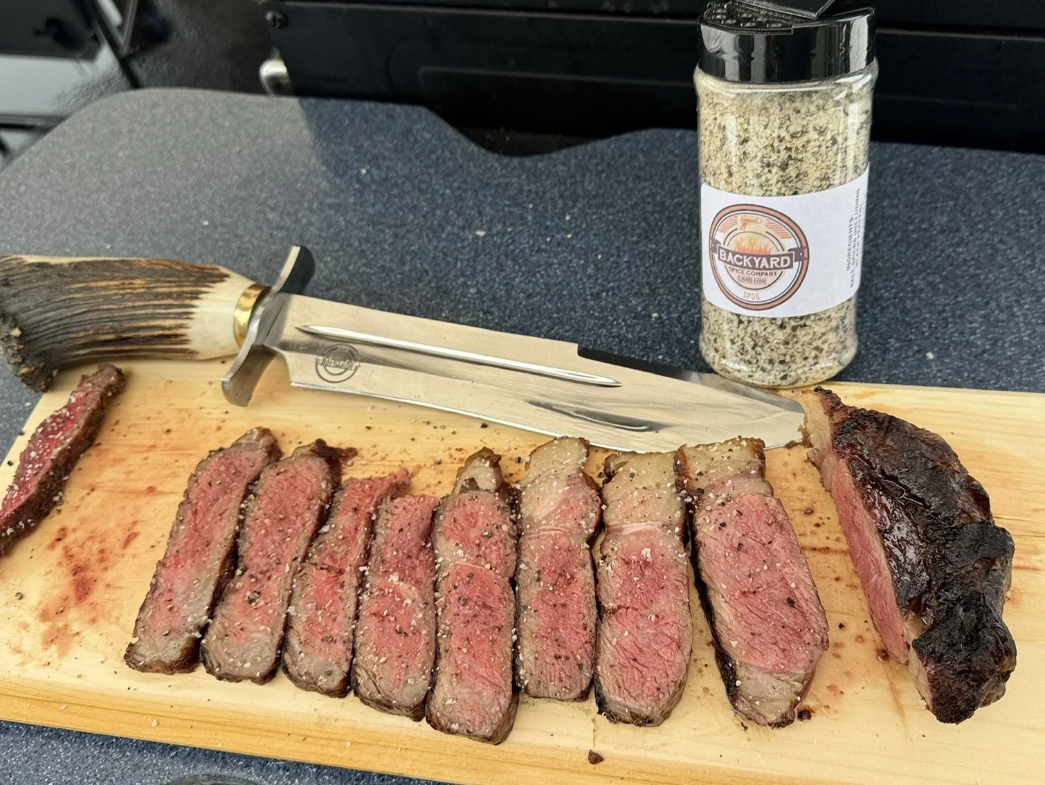Sliced steak seasoned with Backyard Spice Company SPOG blend on a cutting board with the SPOG jar