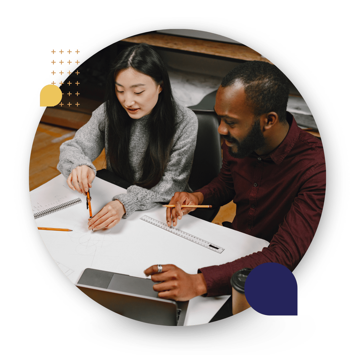 Two coworkers sit together at a table collaborating on a design sketch. One person uses a compass to draw on large paper while the other holds a pencil and ruler. A laptop and coffee cup sit nearby.