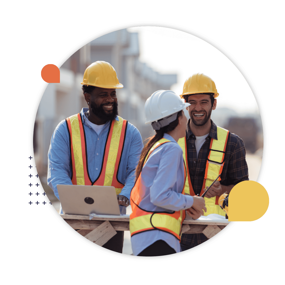 Three construction workers wearing hard hats and high-visibility vests smile and talk together at a worksite, with a laptop resting on a wooden table.