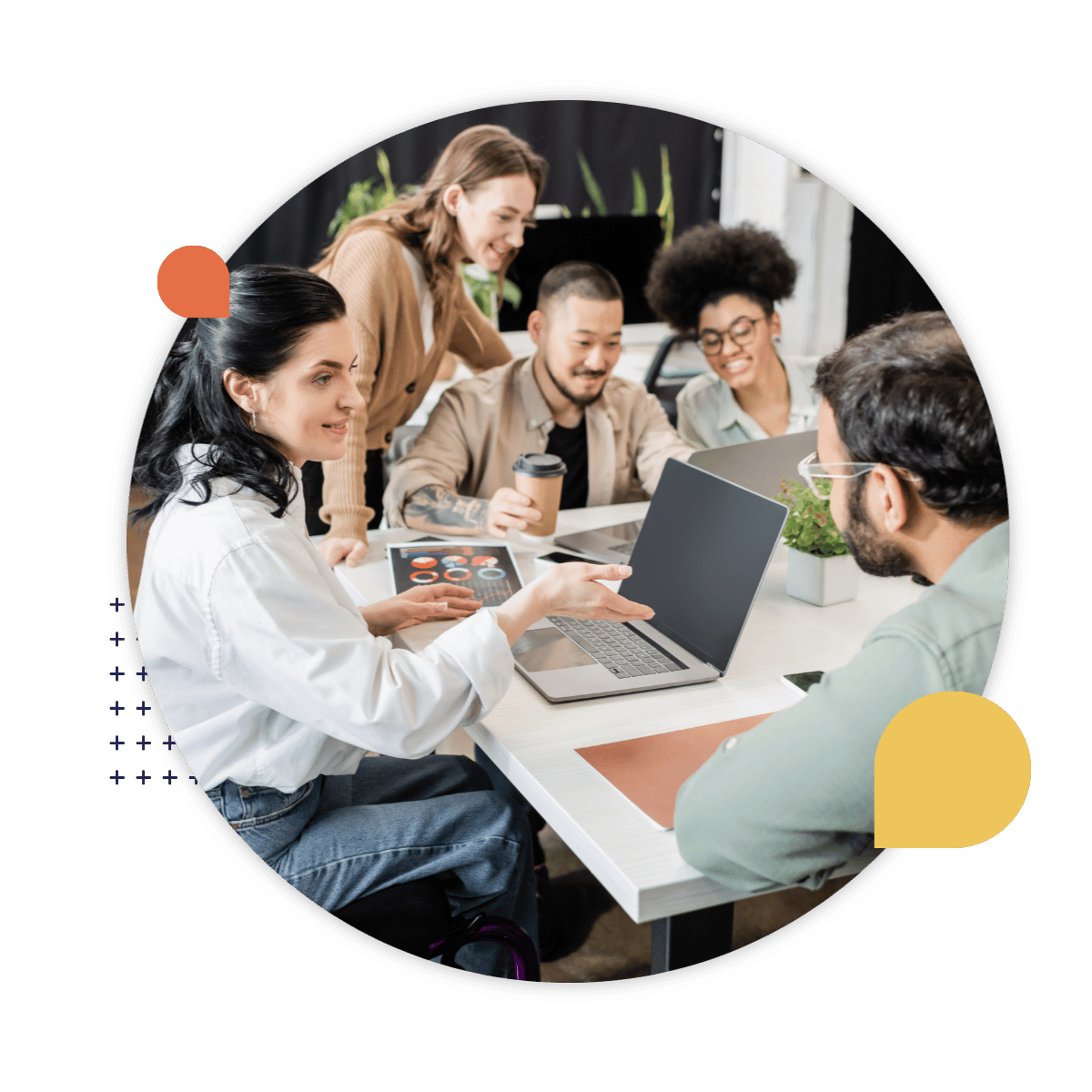 A diverse group of colleagues gathered around a table, talking and looking at a laptop together during a collaborative workplace meeting.
