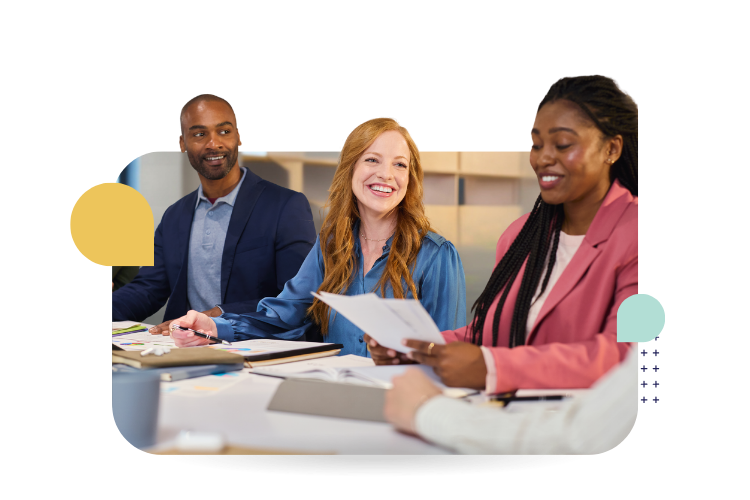Three colleagues smile and collaborate around a table during a workplace discussion, with papers and notebooks in front of them.