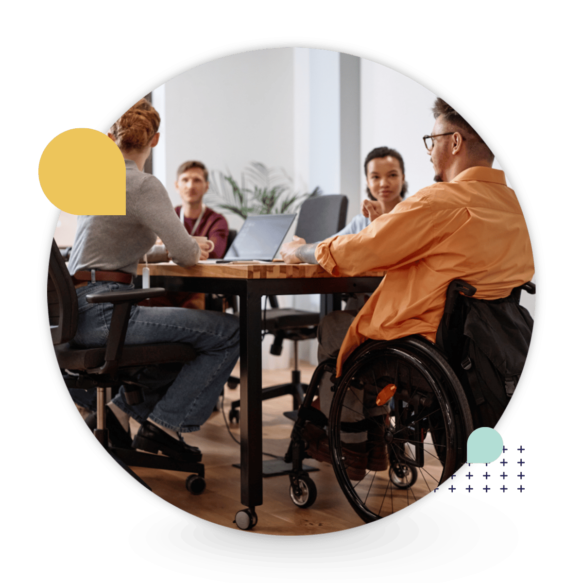 Four colleagues, including a man using a wheelchair, gather around a table with a laptop during a workplace meeting.
