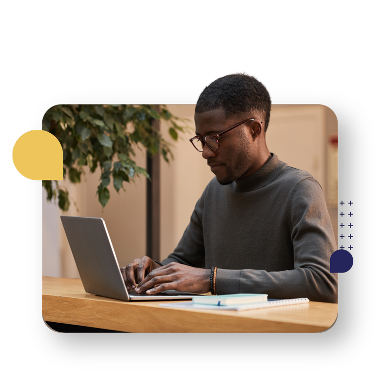 A man wearing glasses works on a laptop at a table, with notebooks nearby.