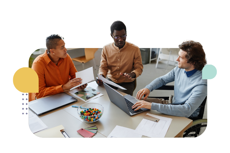 Three colleagues collaborate around a table, reviewing documents and working on laptops in an inclusive office setting.