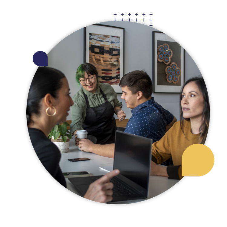 Four Indigenous colleagues gather around a table with laptops, talking and smiling in a collaborative workspace.